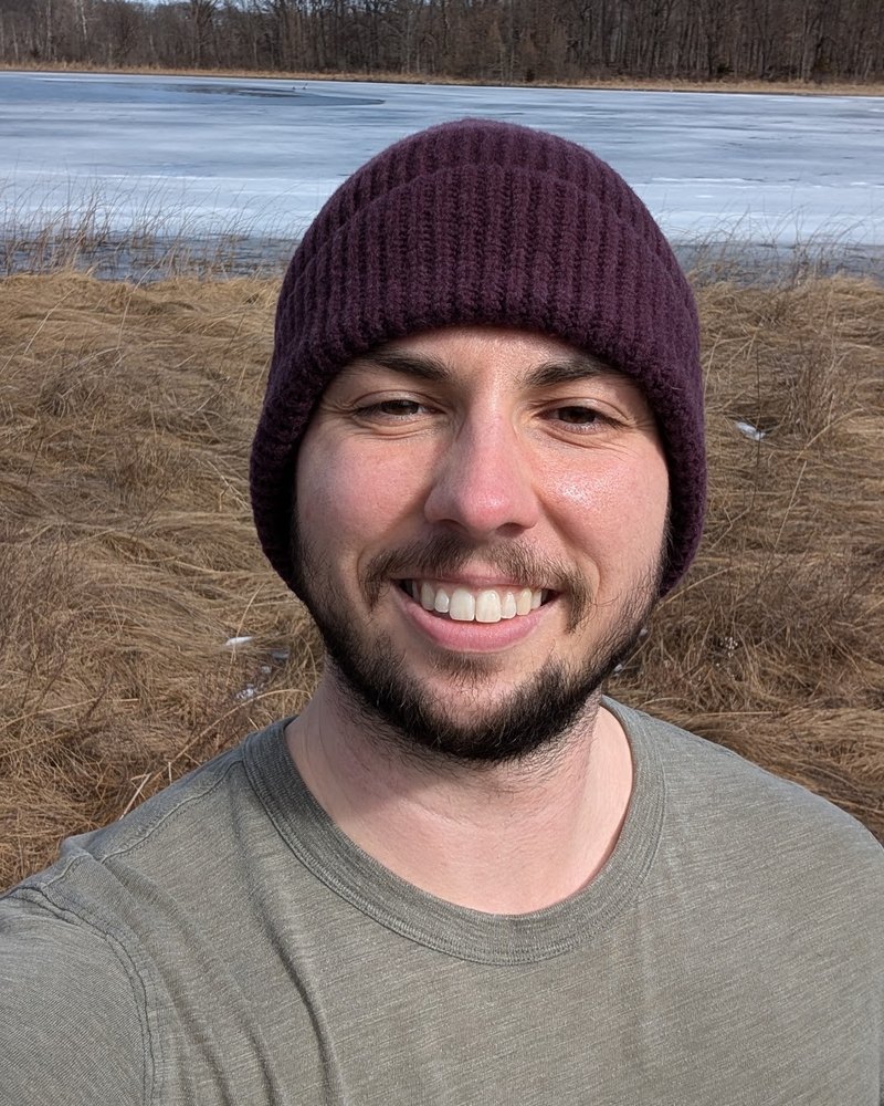 Kalib smiling, wearing a burgundy knit beanie and an olive t-shirt, standing outdoors by a frozen lake and dry grass.